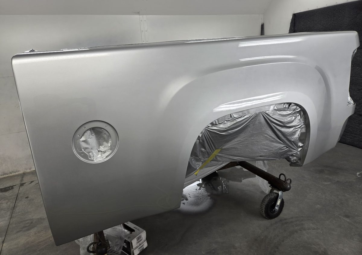 A silver pickup bed side freshly painted and standing on a stand, the wheel behind it wrapped in masking film.