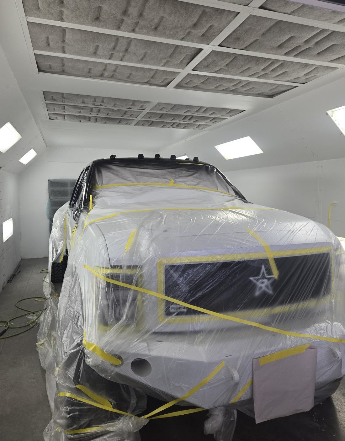A truck fully wrapped in masking film and yellow tape, standing under the lights of the paint booth.