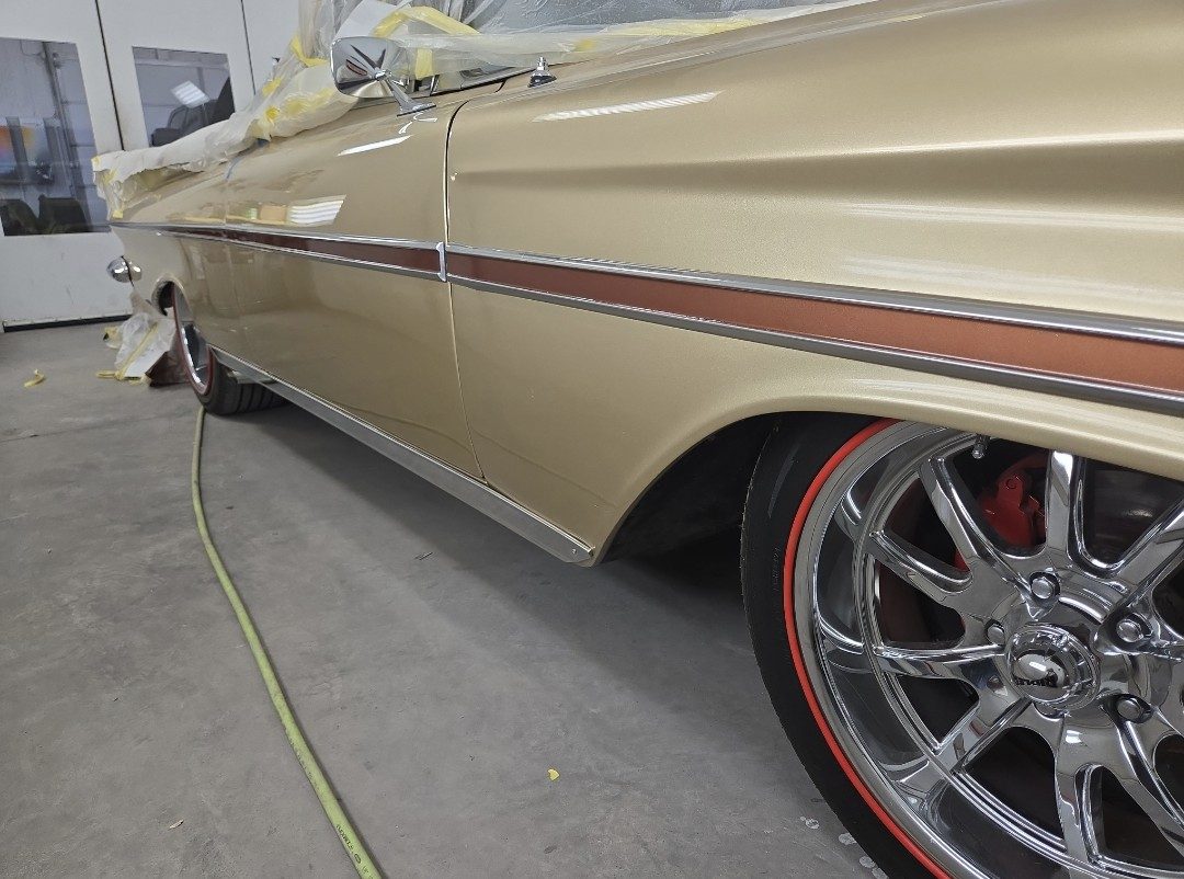 The lower body of a gold classic car with a bronze side stripe, chrome wheels and red line tyres, masking still taped along the top.