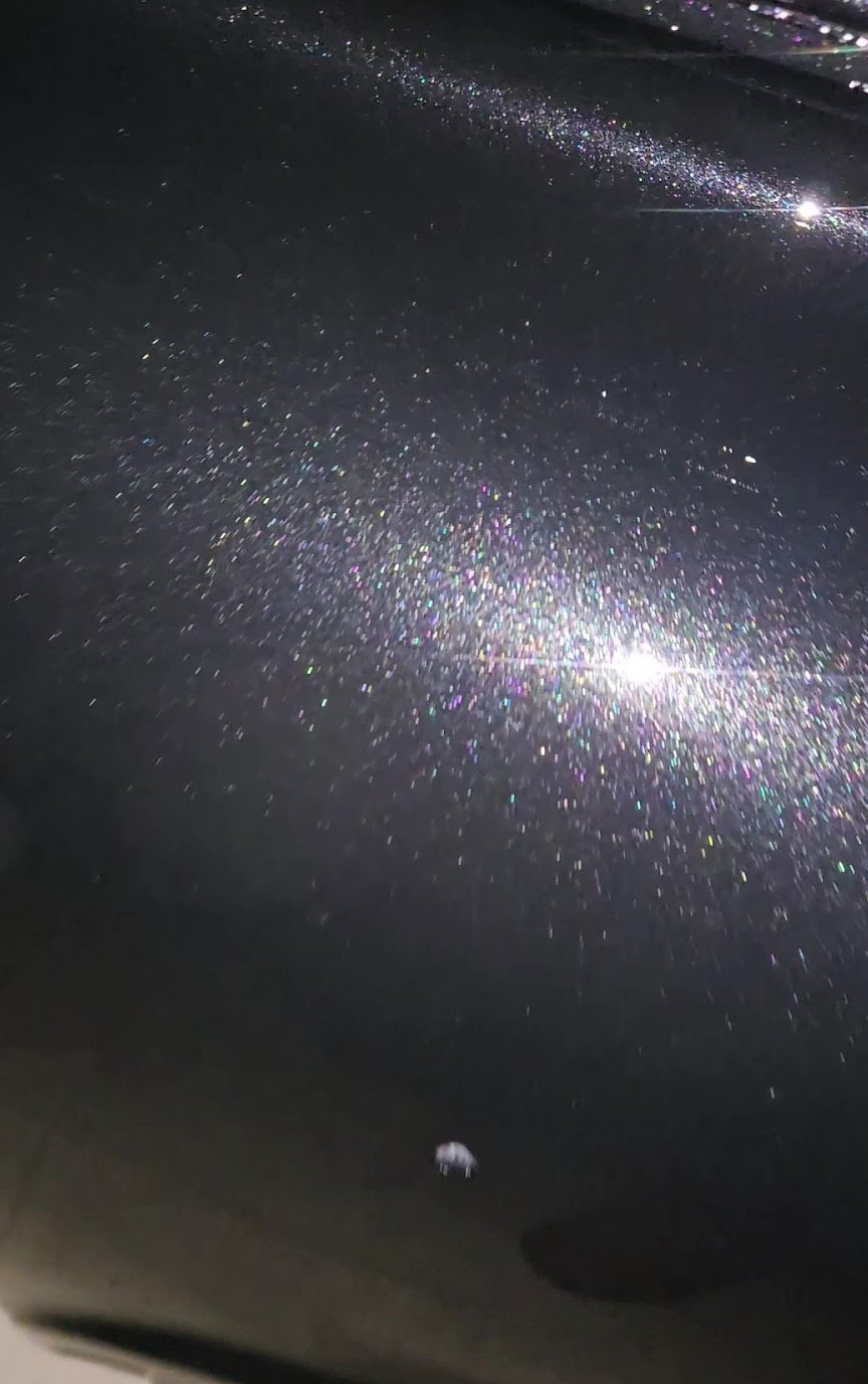 A close view of dark metallic paint, the flake in it catching the light as points of colour.