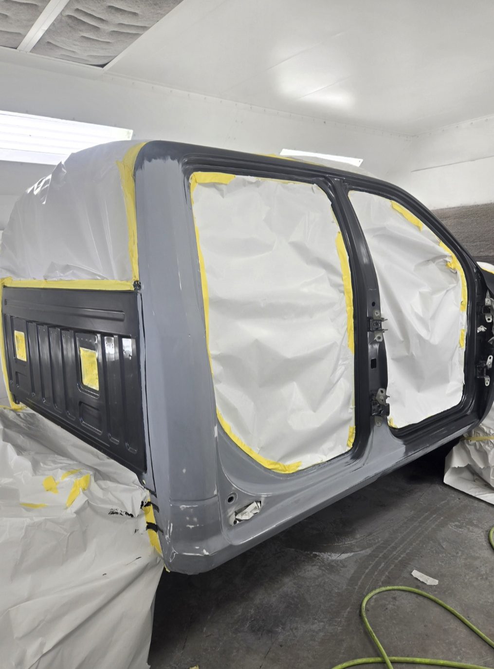 A truck cab in grey primer inside the shop, windows and door openings masked off with paper and yellow tape, ready for colour.
